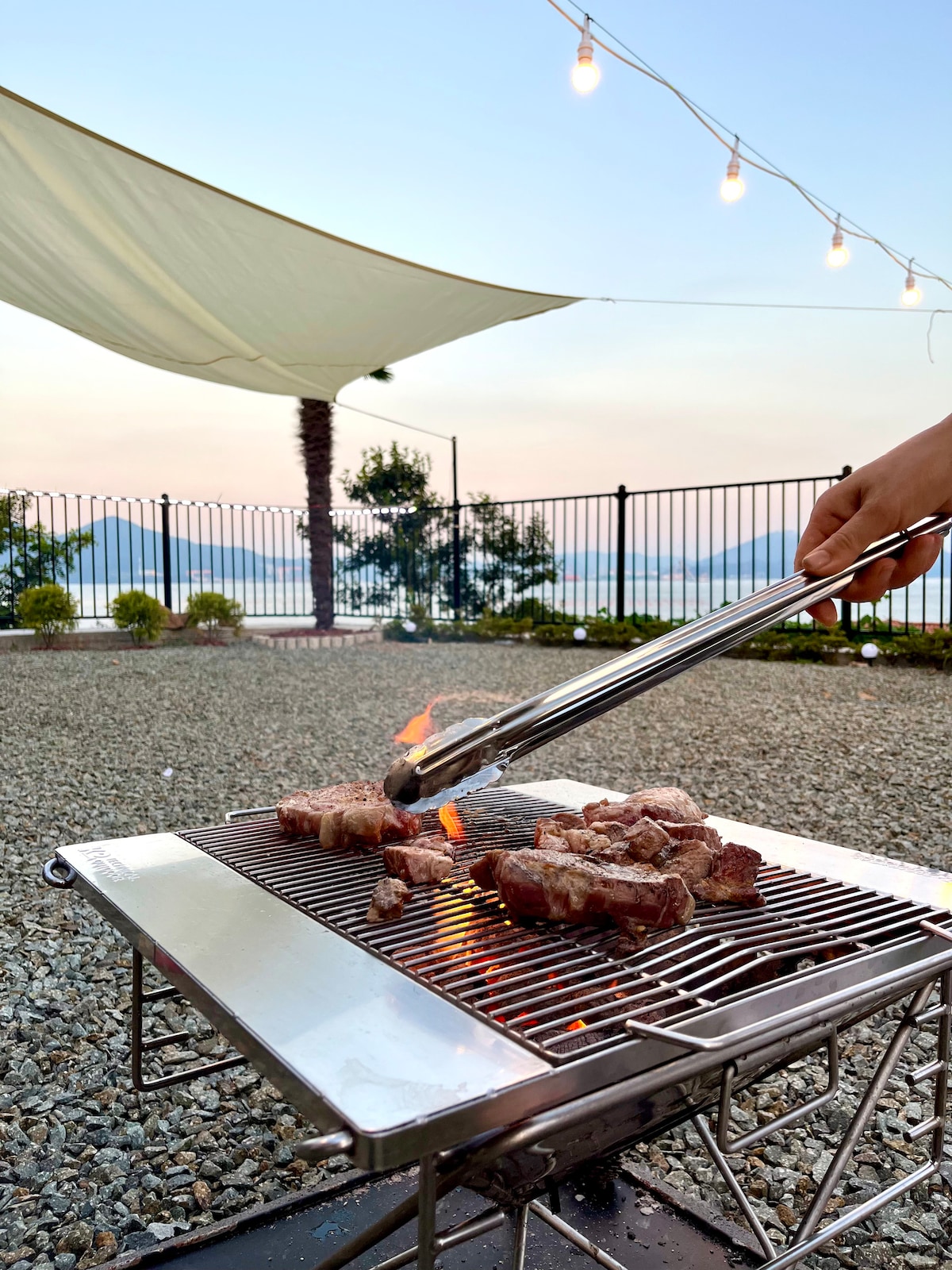 An outdoor barbecue grill is shown with skewered meat being turned over, flames flickering amidst the meat. A spacious gravel area surrounds the grill, and soft lighting from hanging bulbs creates a relaxed setting, with a distant view of the sea and greenery.