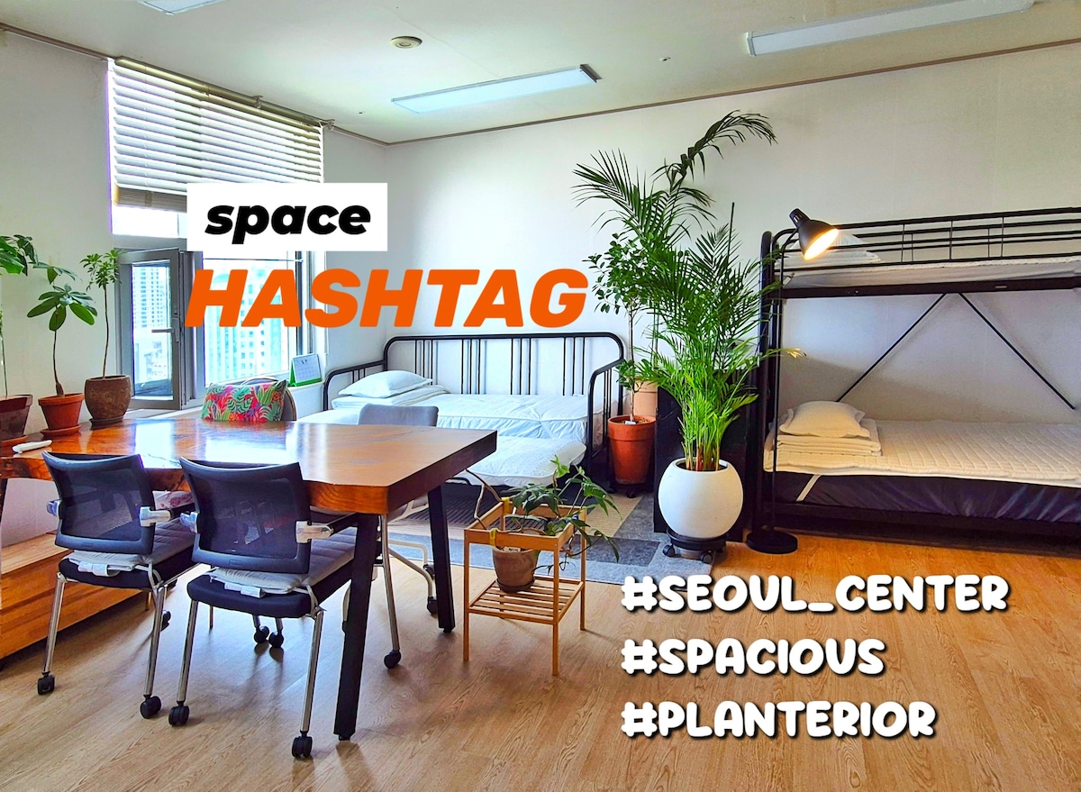 A spacious room features a mix of bedding arrangements, including bunk beds and single beds. A large wooden table with chairs is positioned centrally, surrounded by various plants in pots, enhancing the green decor. Natural light filters through the windows, contributing to a bright environment.