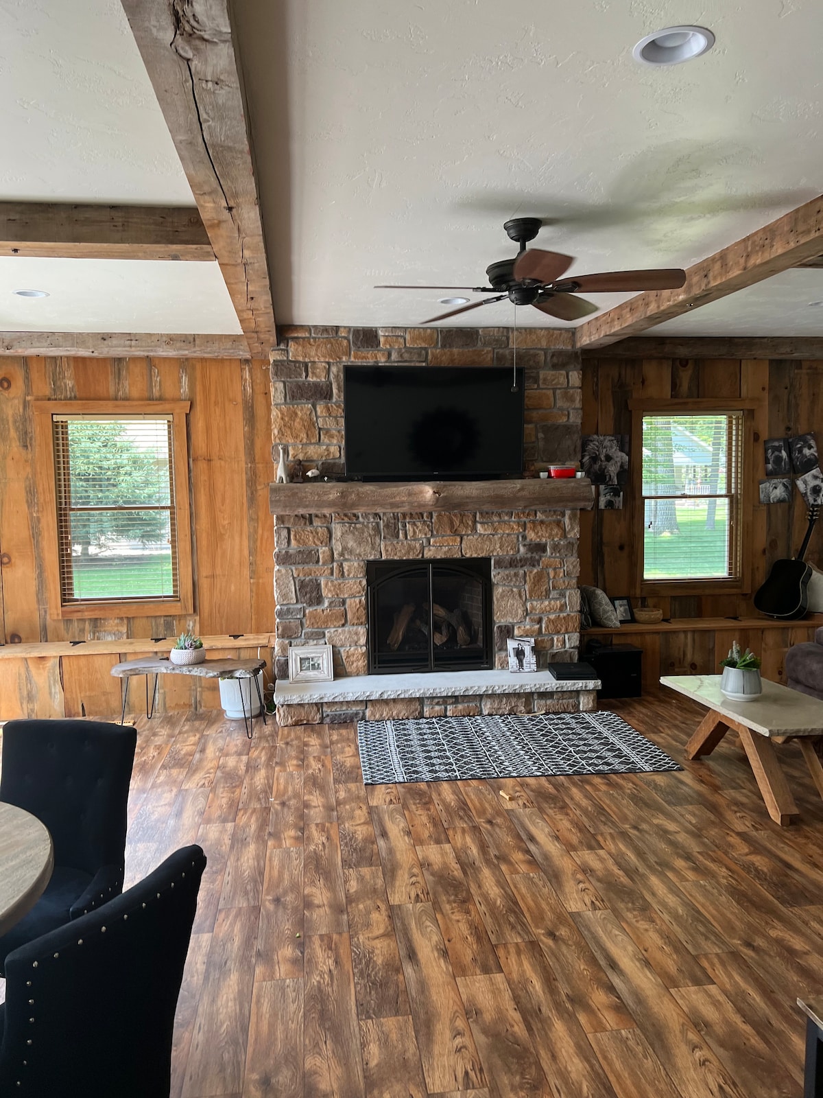 A rustic living area features a stone fireplace as a focal point, with a mounted television above. Wood-paneled walls complement the warm wooden flooring. Natural light filters through the windows, and a black and white patterned rug lies beneath a low coffee table, surrounded by cozy seating.