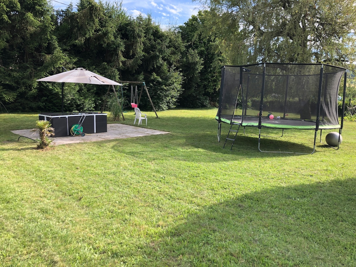 A spacious outdoor area features a grassy lawn surrounded by trees. A shaded seating area with a large umbrella is provided, along with a trampoline and a swing set, creating a family-friendly environment for leisure activities.