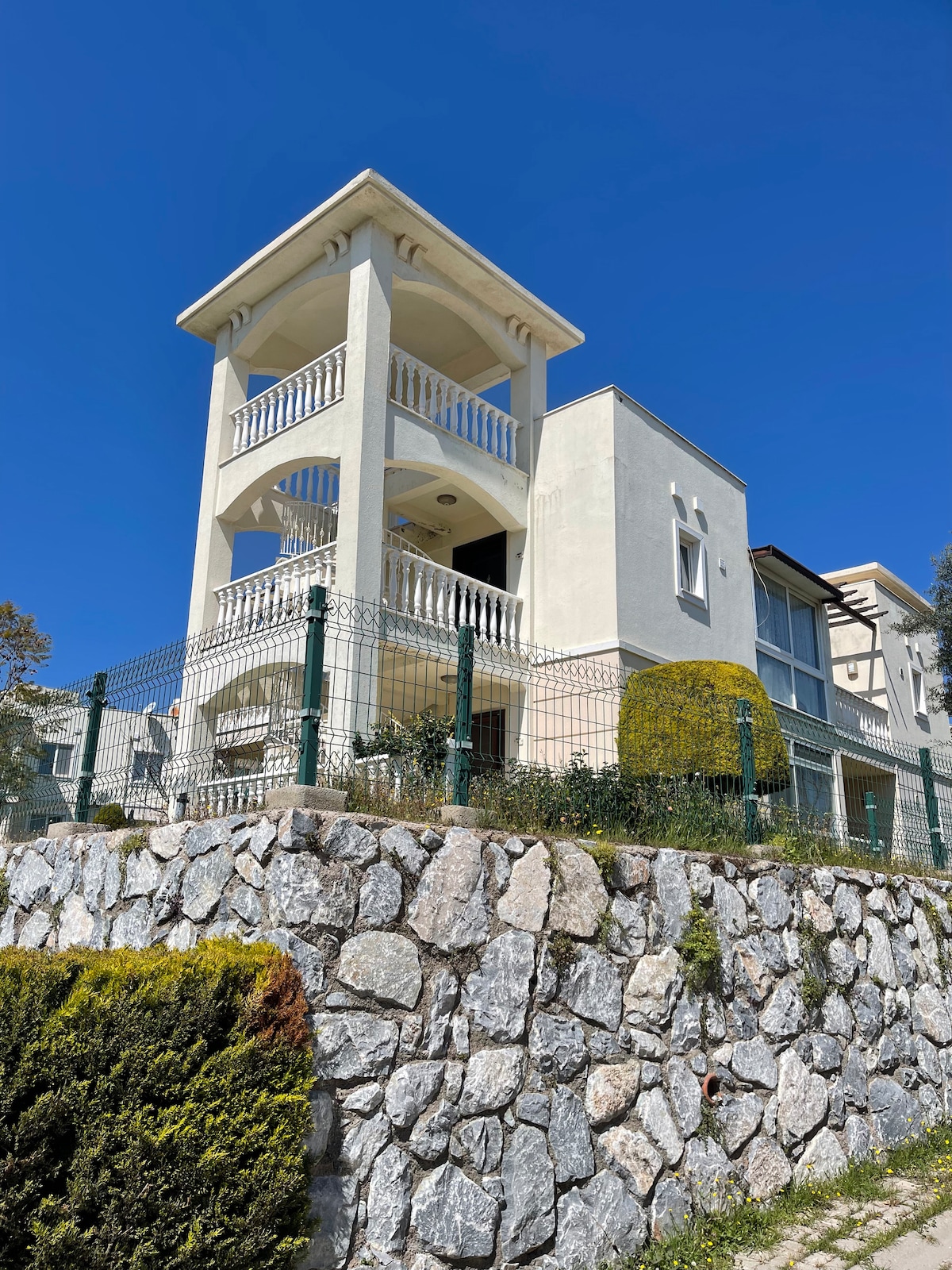 A three-story building is presented, featuring an elegant facade with arched balconies and large windows. Surrounded by a well-maintained stone wall, the property showcases a manicured garden with green hedges, while a clear blue sky is visible above.