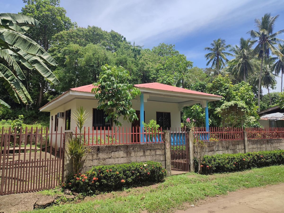 A single-story house is surrounded by lush greenery and palm trees. A low stone wall with a wooden gate encloses the property, while colorful flowers line the front garden. The roof is adorned in terracotta, and a covered porch adds to the welcoming facade.