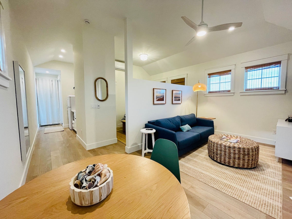 A bright and airy living space is presented, featuring a cozy sitting area with a navy blue couch and round woven coffee table. Natural light filters through the nearby windows, while a dining table garnished with decorative shells adds a touch of charm. A hallway leads toward additional rooms.