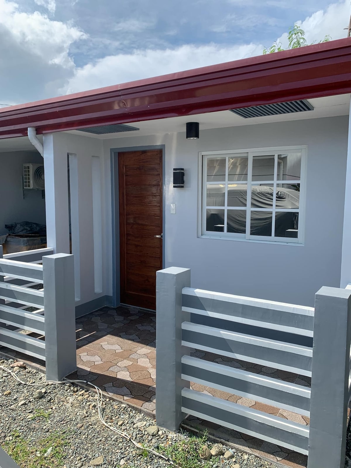 A newly constructed exterior is shown, featuring a welcoming wooden door and a small front porch area. A light fixture is installed by the entrance, while a window provides natural light. The gray fencing adds a modern touch to the overall design.
