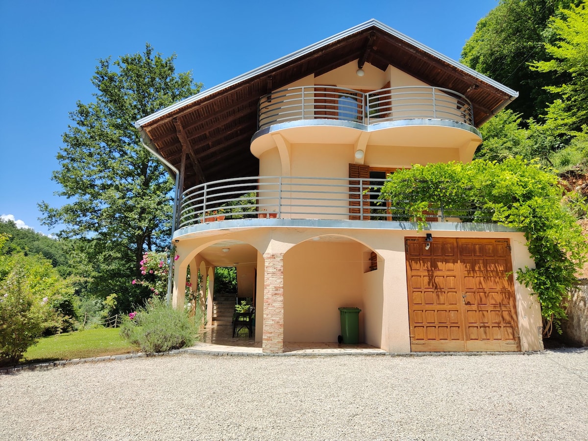 The villa features a two-story structure with a wraparound balcony, surrounded by lush greenery. The exterior is complemented by stone and wooden accents, and a spacious gravel driveway leads up to the entrance, providing access to the garage area.