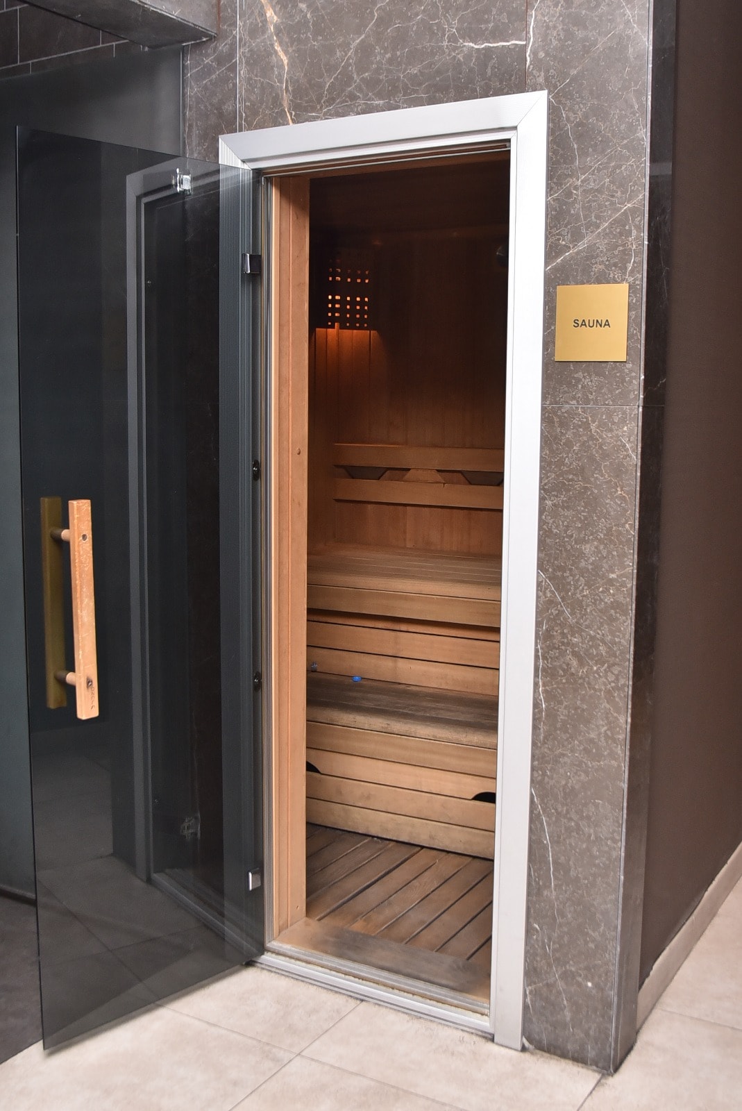 A modern sauna is framed with sleek glass doors and features wooden benches. Natural wood accents provide a warm atmosphere, while LED lighting adds a contemporary touch. The sauna's entry is marked with a simple sign indicating 'SAUNA'.