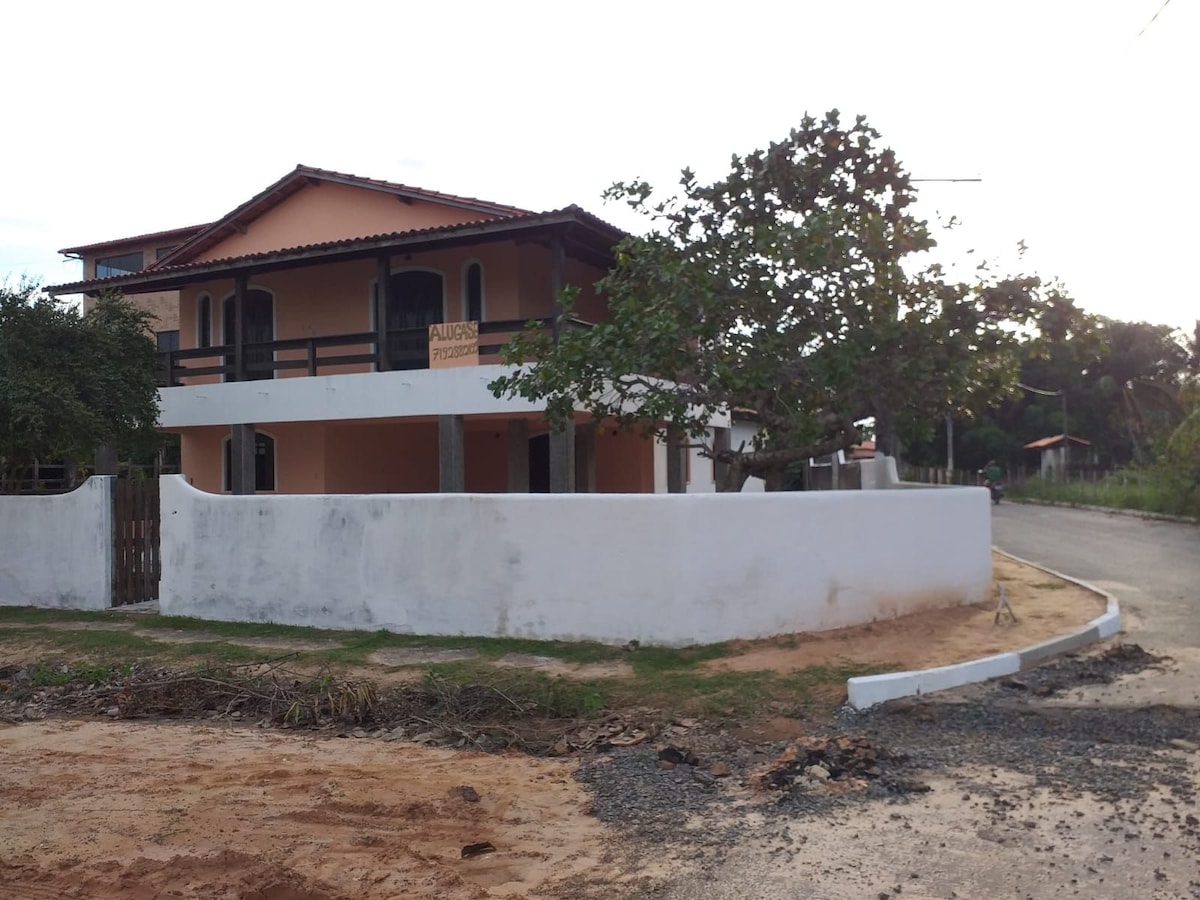 A spacious two-story house is featured in the image, displaying a light-colored exterior with a terracotta roof. Surrounding greenery is visible, along with a partially enclosed yard and a path leading to the road.
