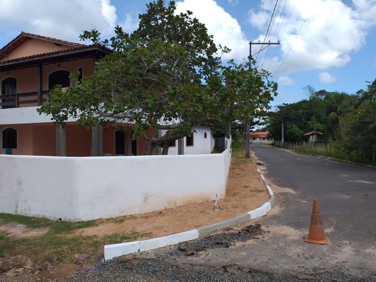 The exterior of a spacious house is depicted, featuring a large tree providing shade near the entrance. A partially visible white wall surrounds the property, and the paved road leads into the distance. The scene is complemented by a blue sky with scattered clouds.