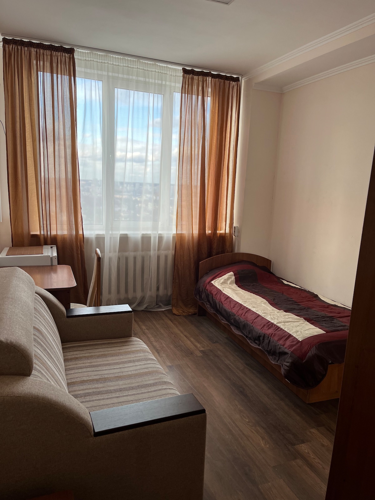 A cozy single room is presented with a spacious bed covered in a maroon and beige quilt. A comfortable armchair is positioned next to the window, which is dressed in flowing brown and sheer curtains. Natural light fills the space, enhancing the warm tones of the decor.