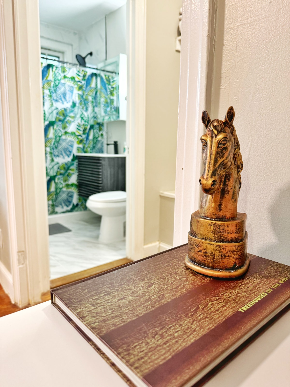 A decorative horse statue sits atop a wooden book on a tabletop, with a glimpse of the bathroom visible in the background. The bathroom features a colorful shower curtain and modern fixtures, creating a lively contrast against the neutral tones of the hallway.