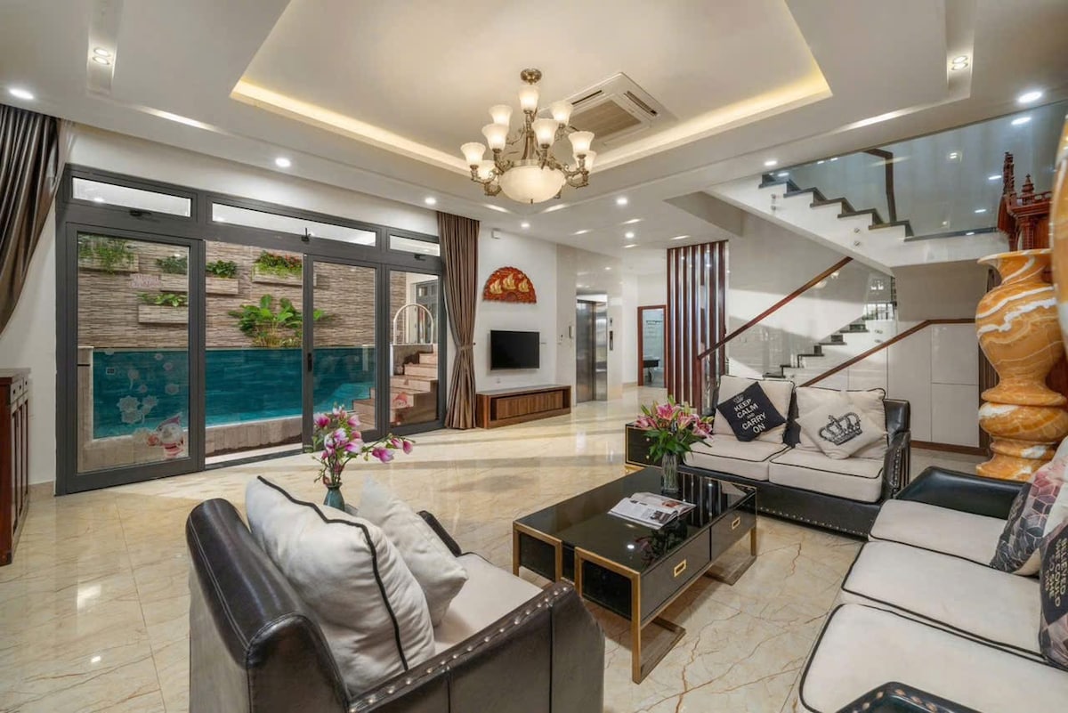 An elegantly furnished living area features plush sofas and a coffee table, complemented by decorative pillows. Floor-to-ceiling windows offer views of an outdoor space with a private pool. A staircase and modern decor elements are visible, enhancing the open layout.