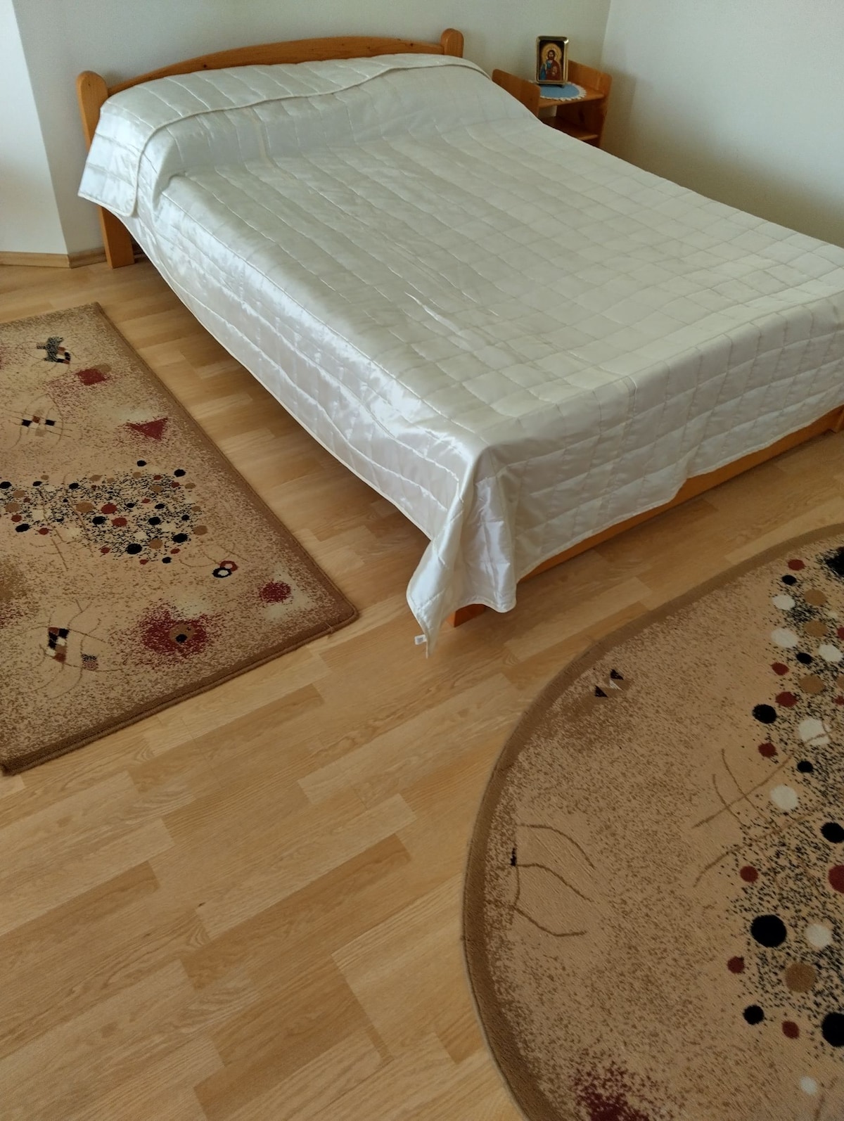 A spacious bed covered with a shiny white quilt is positioned on a light wooden floor. Two area rugs with abstract patterns in warm tones are present, enhancing the warmth of the room.