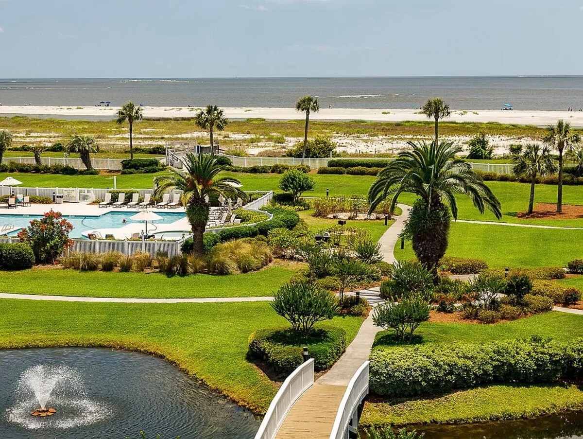 The image captures the lush landscaped grounds of the St. Simons Grand, featuring a swimming pool, palm trees, and a serene lagoon with a fountain. The beach can be seen in the distance, alongside the peaceful ocean, creating a relaxing outdoor environment.