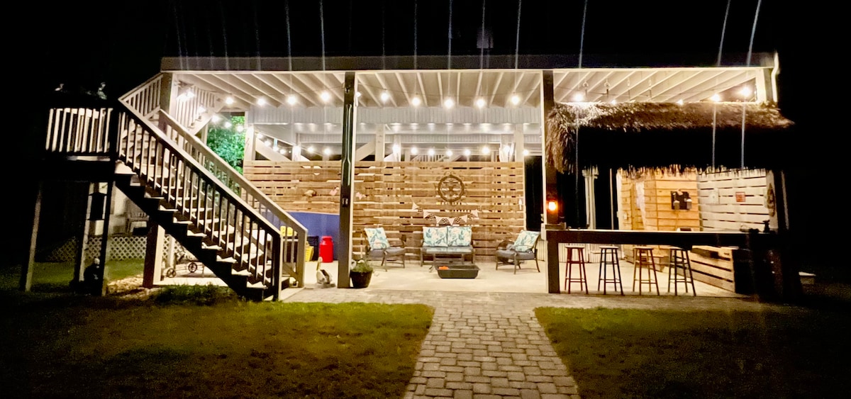 An outdoor living area is illuminated under evening lights, featuring wooden walls and a seating arrangement with blue and white patterned cushions. A staircase leads to the upper level, with a fire pit and tiki bar visible in the background, enhancing the inviting outdoor atmosphere.
