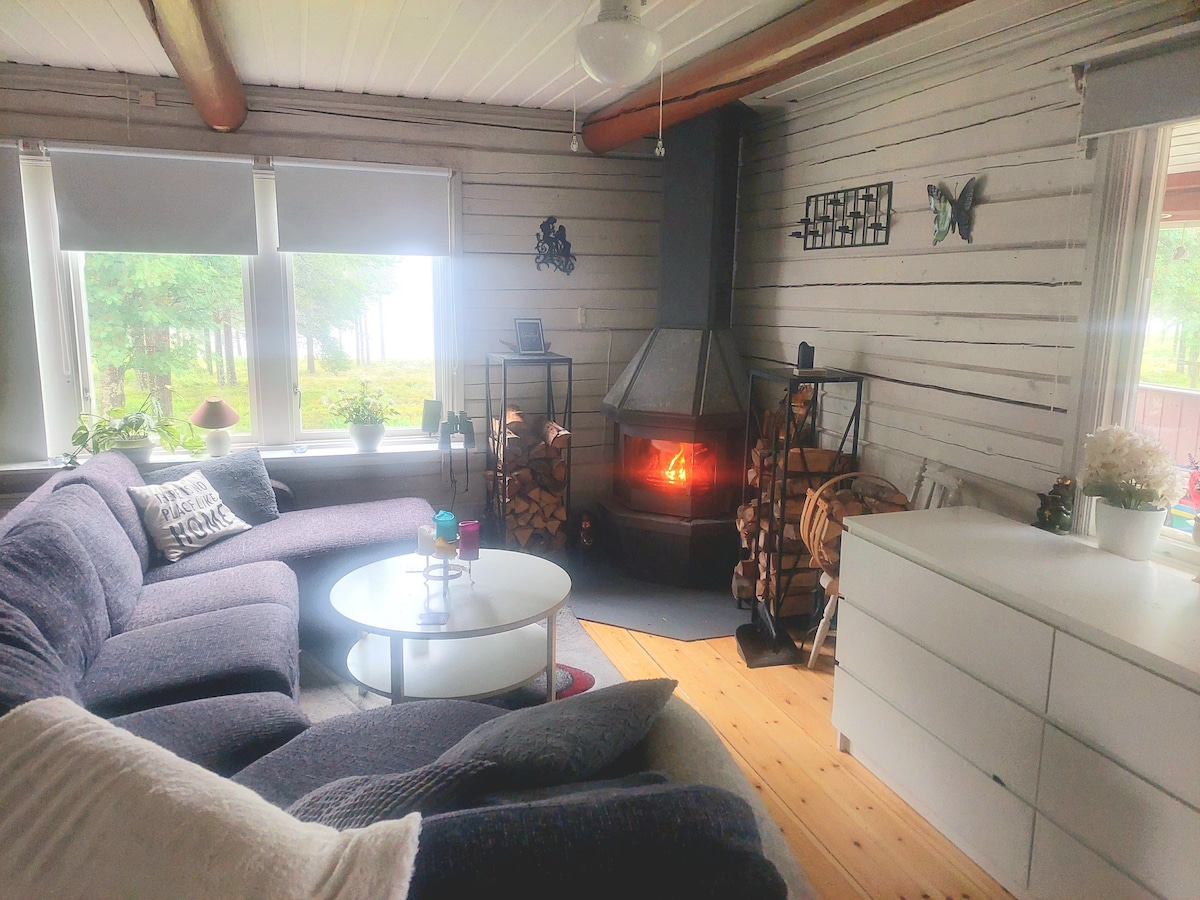 A cozy living area is presented with a dark sectional sofa and a round coffee table at the center. A modern fireplace is situated against a wood-paneled wall, creating a focal point. Natural light filters in through window shades, illuminating the room's inviting features.