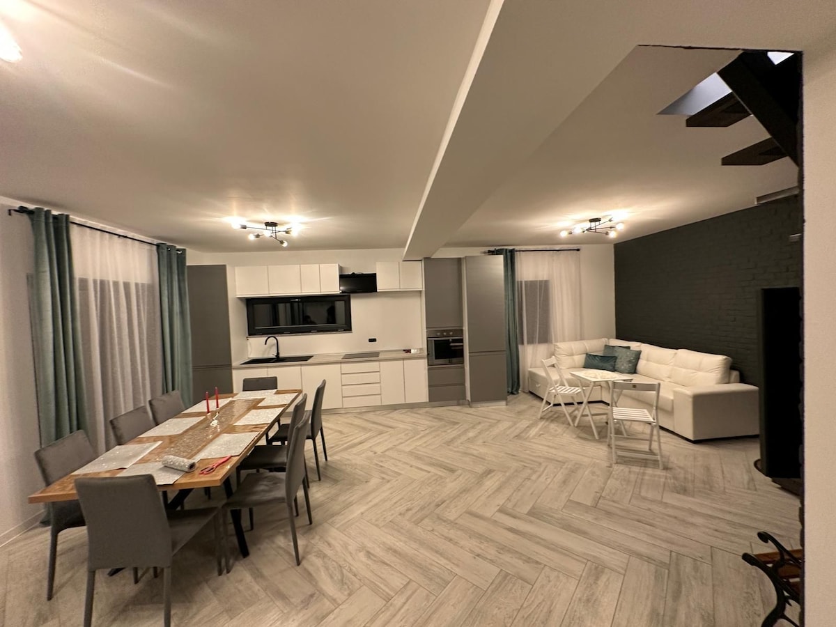 An open-concept living space is highlighted, featuring a modern kitchen with sleek cabinetry and a dining table surrounded by multiple chairs. A comfortable sectional sofa with light-colored cushions is situated in the lounge area, complemented by large windows dressed with soft drapes.