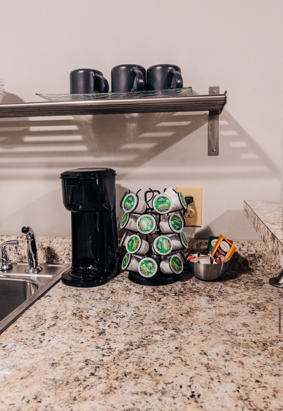 A compact kitchen counter features a coffee maker next to a rotating stand filled with coffee pods. Above, a shelf holds several dark mugs. A small bowl with assorted sugar and cream packets rests beside the sink, which is integrated into the granite countertop.
