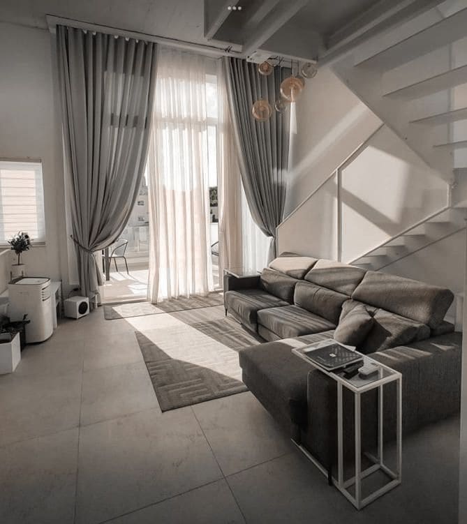 A light-filled living area features large windows draped with sheer curtains, allowing natural light to illuminate the space. A comfortable sofa is complemented by a minimalist side table. A staircase leads to the upper level, enhancing the open and airy atmosphere.