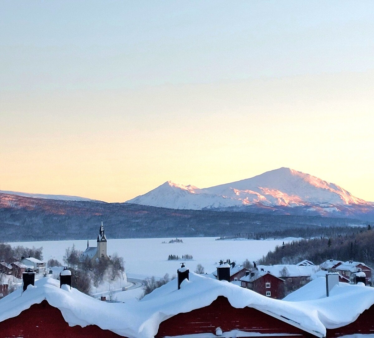 A serene winter landscape is portrayed, featuring snow-covered rooftops in the foreground. Beyond, a tranquil lake reflects the soft hues of sunrise, framed by majestic mountains in the distance. A church steeple is visible among the snow-laden trees.
