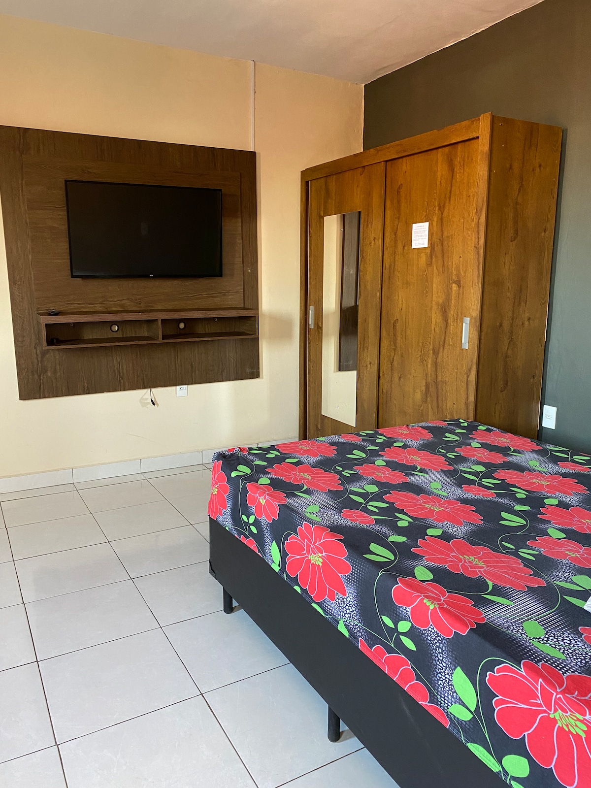 A cozy sleeping area features a spacious bed adorned with a floral patterned cover. A wooden wardrobe is positioned beside the bed, while a wall-mounted television is installed above a small shelf. The room is illuminated by natural light from a nearby window.
