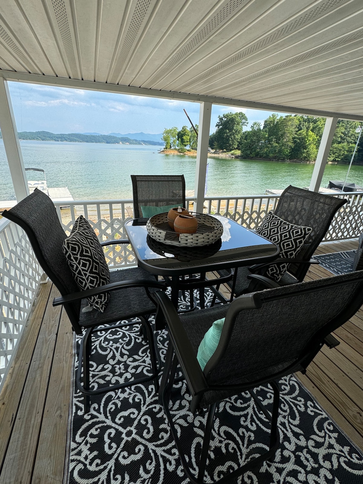 A dining area on the porch includes a table surrounded by four chairs, all positioned on a patterned area rug. A bowl of decorative items sits at the center of the table. The serene lake view and distant greenery enhance the setting.