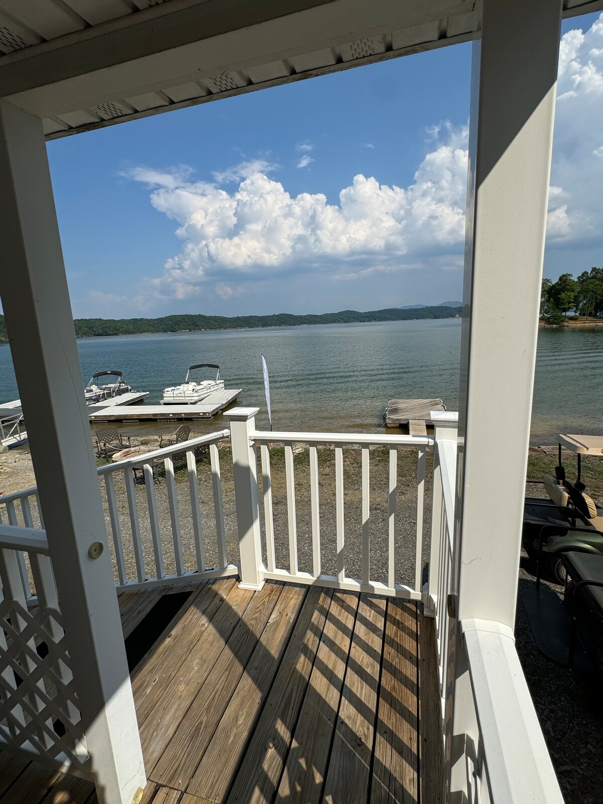The image captures a view of the lake from a porch, showcasing calm water under a partly cloudy sky. Wooden decking extends outward, with boats visible along the shore. The white railing frames the scene, inviting a sense of tranquility.