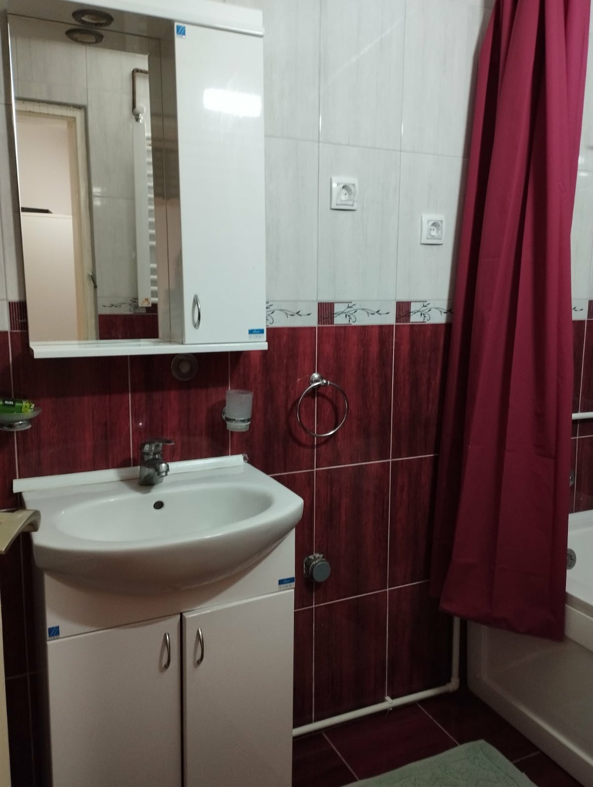 A functional bathroom is displayed, featuring white cabinetry and a matching sink. A mirrored cabinet is mounted above the sink. A red shower curtain hangs beside a bathtub, with white tiles creating a clean and modern ambiance.