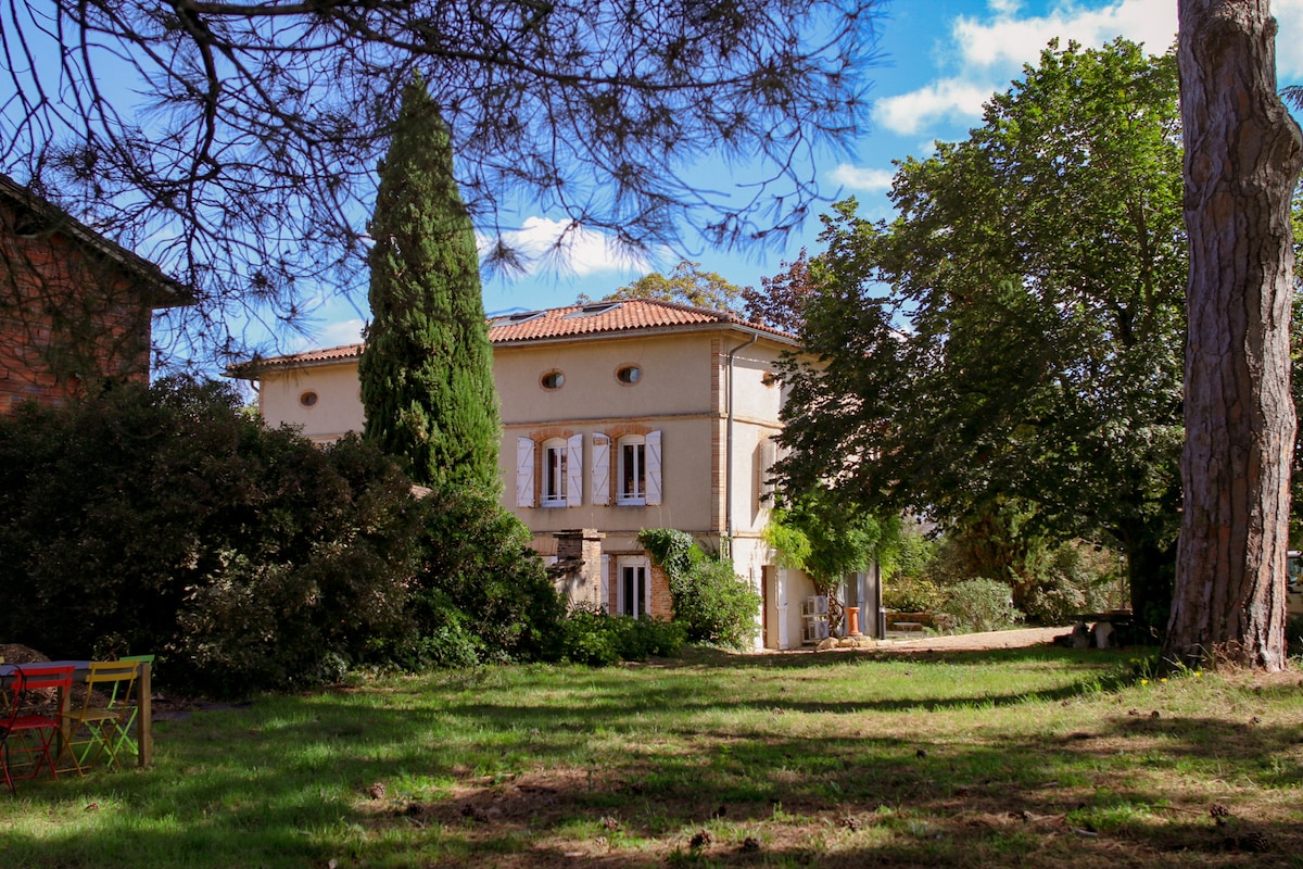 A charming house is set in a lush green landscape, framed by diverse trees and shrubs. Sunlight filters through the leaves, creating a tranquil atmosphere. The exterior features white windows and a terracotta-tiled roof, while a spacious lawn invites relaxation.