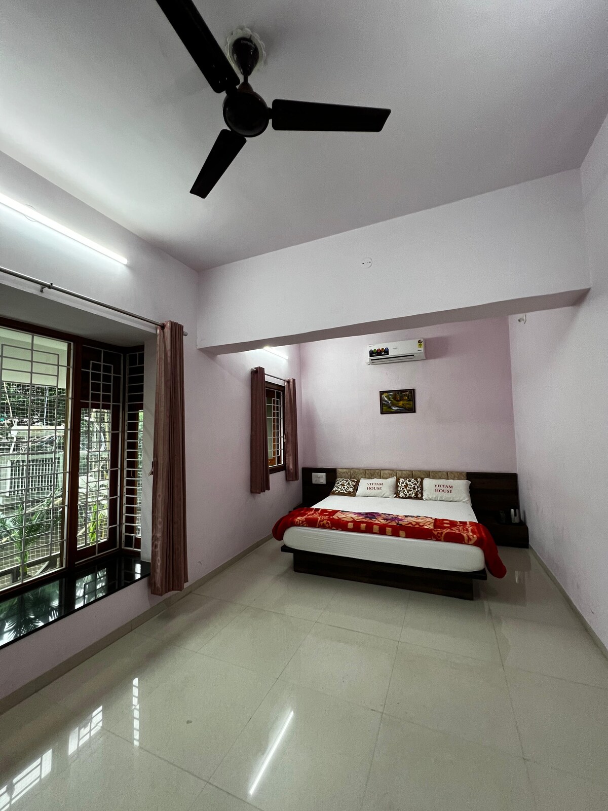 A well-lit bedroom features a spacious bed with decorative pillows and a patterned blanket. Large windows allow natural light to illuminate the room, revealing a view of the greenery outside. A ceiling fan is also present, enhancing comfort.