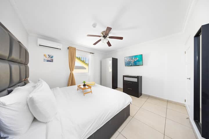 غرفة نوم مكيفة الهواء وواسعة مع تلفزيون بشاشة مسطحة ومروحة سقف وستائر تعتيم وبعض التخزين.