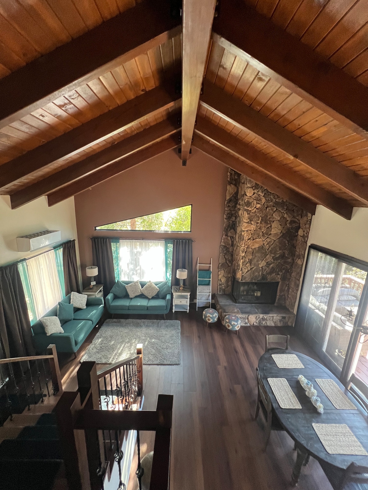 The interior space features high, wooden ceilings and a large stone fireplace. A teal sectional sofa is gathered around a plush area rug, while natural light streams through large windows, illuminating the cozy dining area with a long table set against the backdrop of the living room.