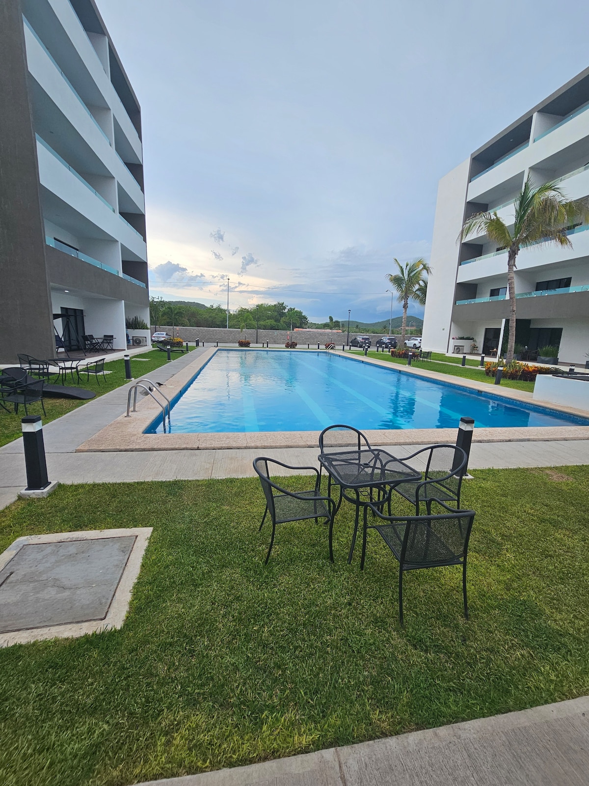 A clear view of the outdoor swimming pool is presented, surrounded by well-maintained grass and palm trees. Several black metal chairs and tables are arranged nearby, providing a relaxing space for guests. Two modern buildings flank the pool area, enhancing the open atmosphere.