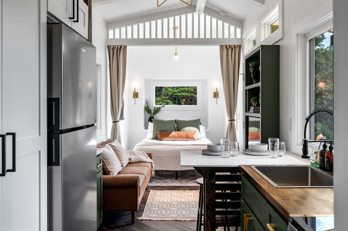 The interior showcases an open layout with a compact kitchen on the left and a cozy living area featuring a sofa. Natural light enters through large windows, illuminating the inviting sleeping area, which is adorned with green accents and soft bedding.