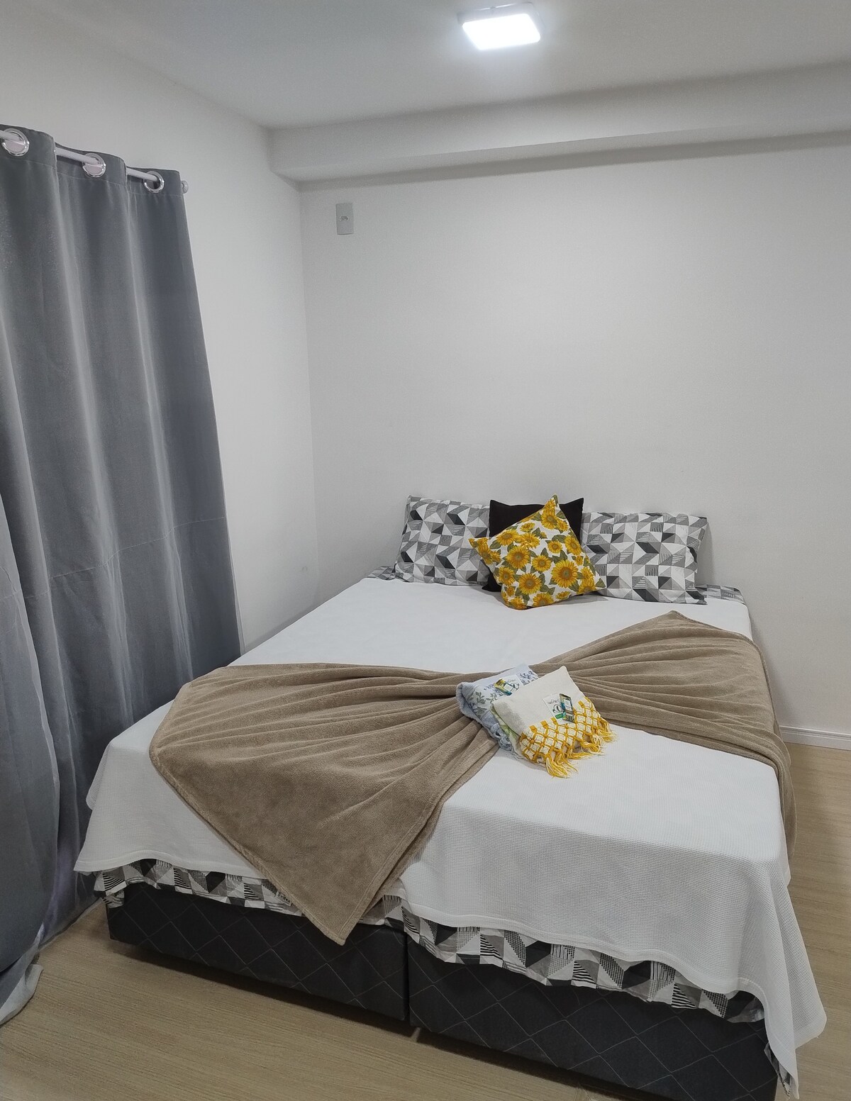 A cozy sleeping area is arranged with a spacious bed layered with a beige blanket and two patterned pillows. Decorative cushions add color, while light gray curtains frame the window. The room is brightly lit and features neutral-colored walls.
