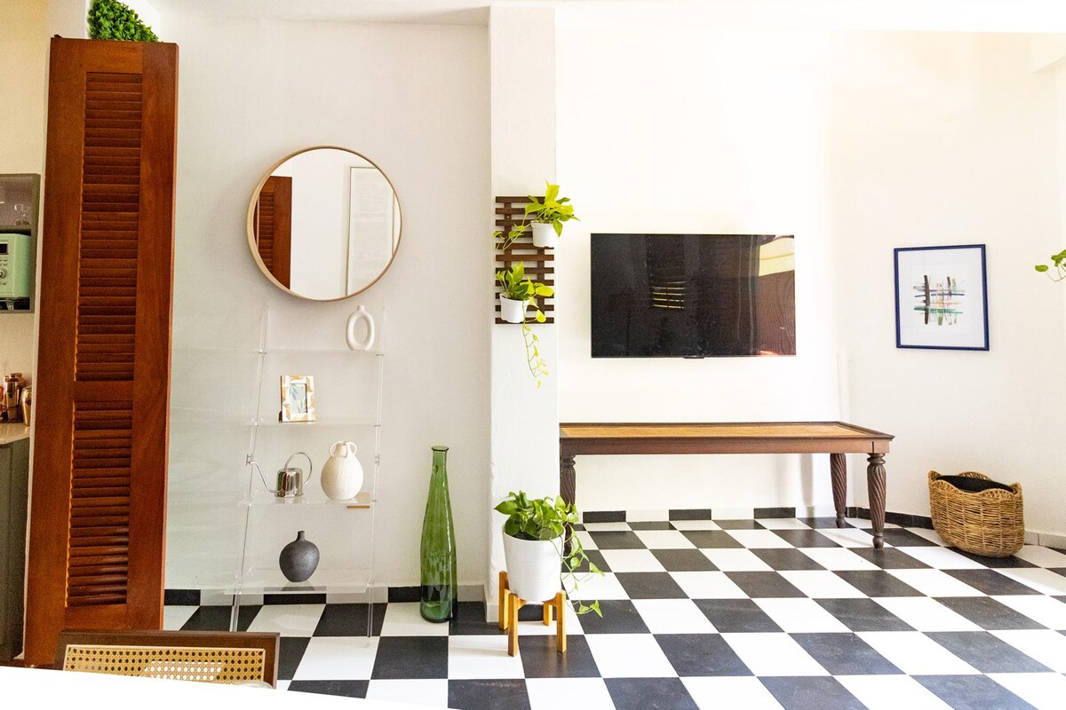 A living area showcases classic black and white checkered floors, complemented by a wall-mounted television and a decorative mirror. Minimalist shelves display selected decor items alongside a tall green vase. A wooden table and a woven basket add functional elements to the space.