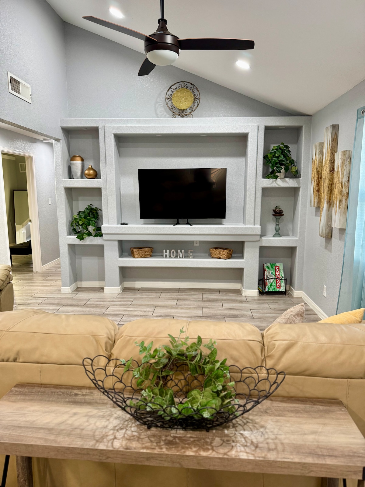 A spacious living area features a comfortable beige couch in the foreground, accompanied by a decorative table with a plant display. A wall-mounted television is framed by a built-in shelf, adorned with plants and decorative items. Soft light enters through large windows, enhancing the calming atmosphere.