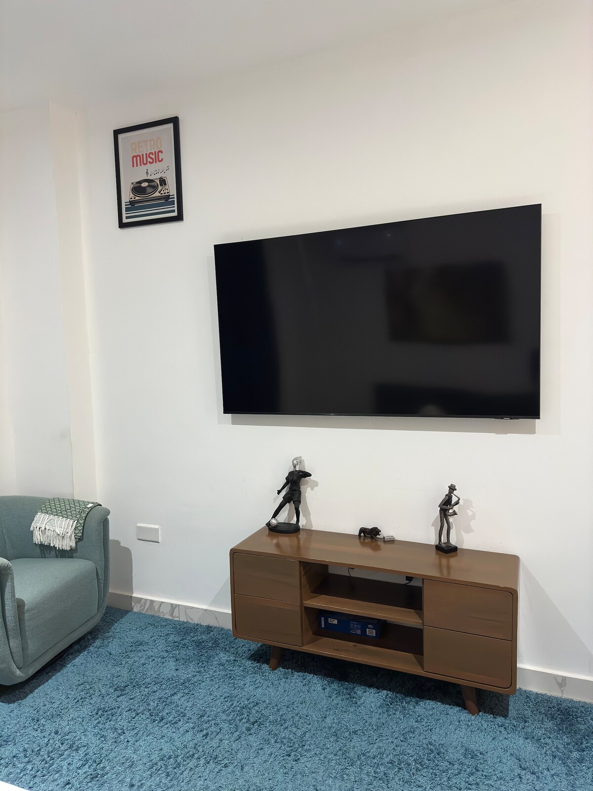 A modern living area features a flat-screen television mounted on a white wall above a stylish wooden console. Two decorative figurines are displayed on the console, while a comfortable green armchair is positioned next to a soft blue area rug.