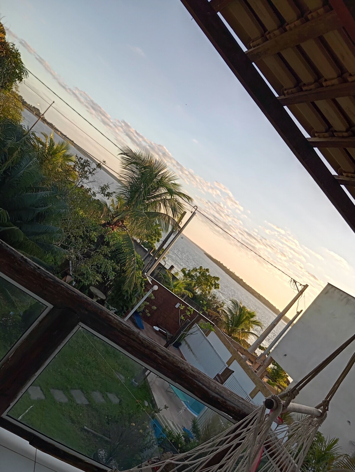 A balcony view offers a serene landscape featuring lush palm trees and a calm waterfront. The sunset casts warm hues across the sky, enhancing the tranquil atmosphere. A hammock is visible, inviting relaxation while enjoying the picturesque surroundings.