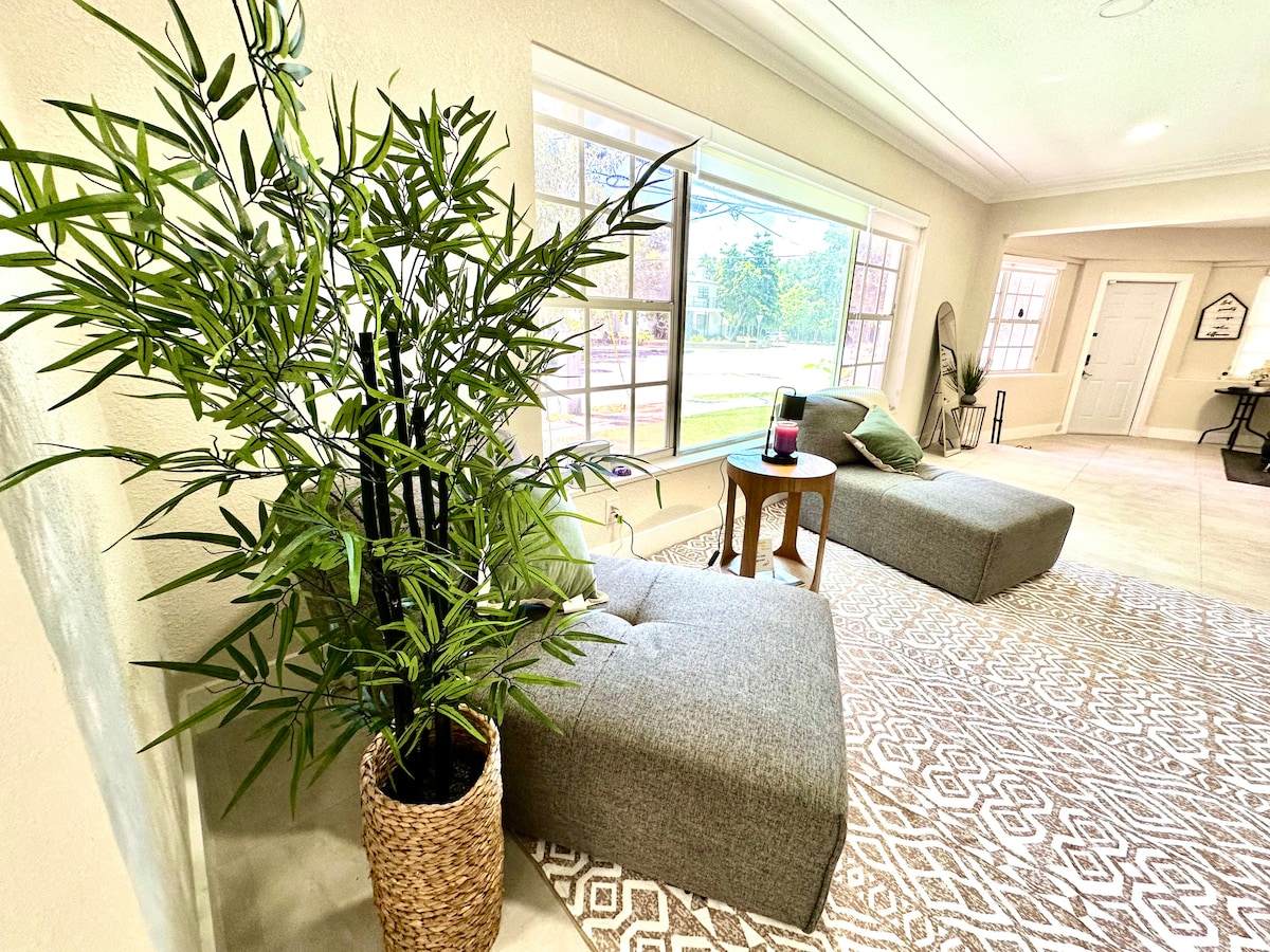 A shared living space features two gray lounge chairs positioned near a large window, allowing natural light to fill the room. A small wooden side table is placed between the chairs, complemented by a decorative potted plant nearby, creating a relaxed environment.
