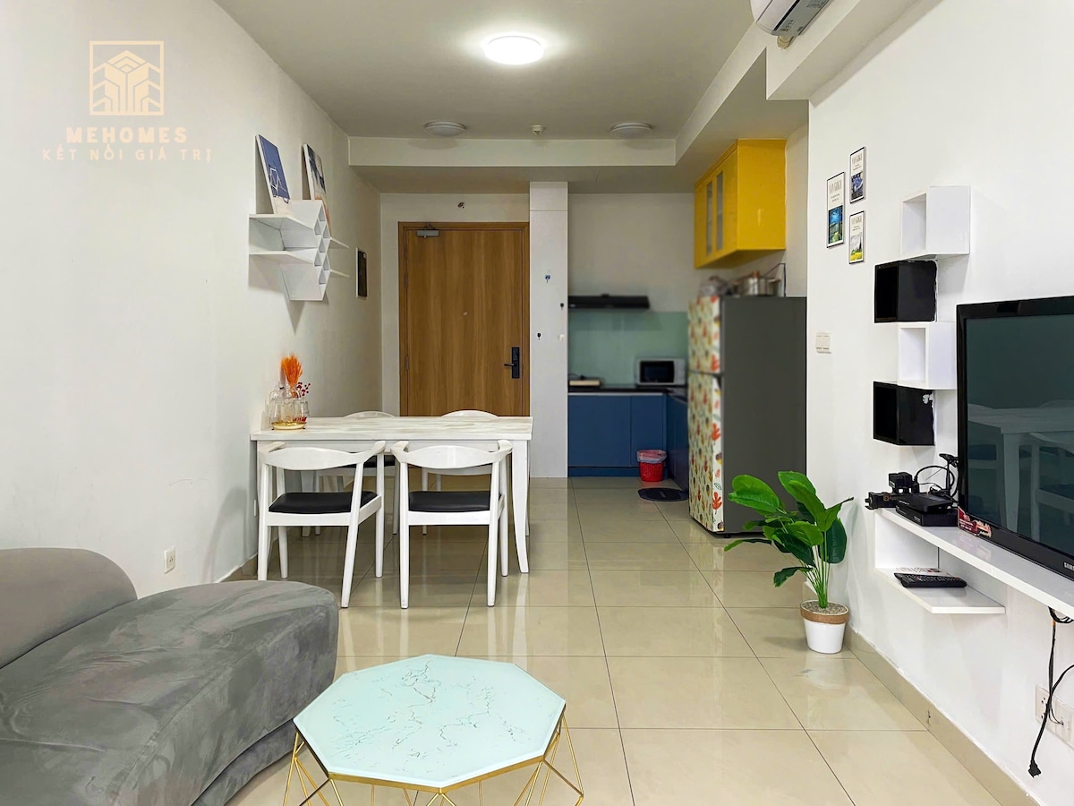 An open living space features a bright kitchen area in blue and yellow cabinets. A light dining table with white chairs is visible, complemented by a small turquoise coffee table. A green plant adds a fresh touch, while ample natural light fills the room.