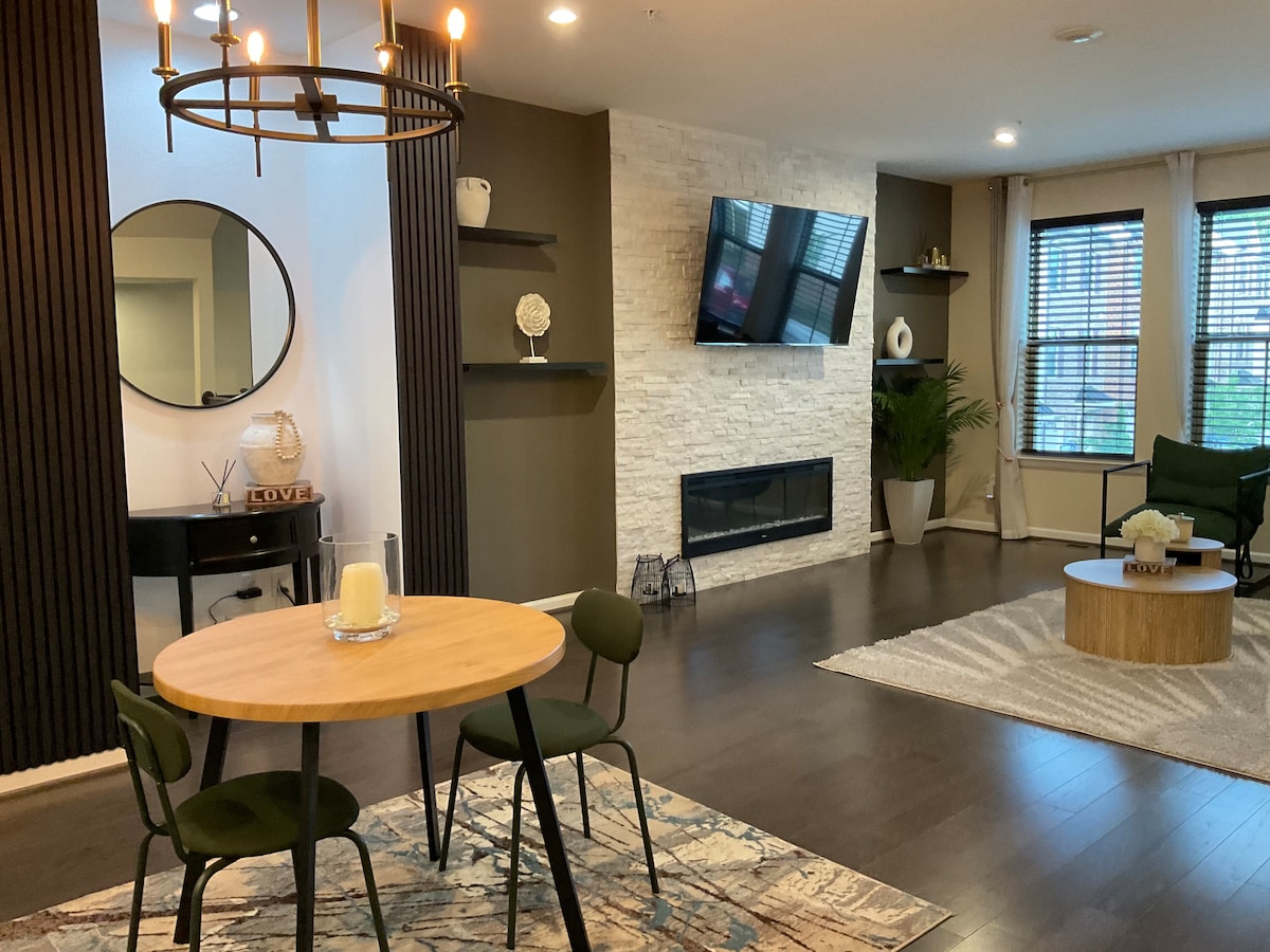 A contemporary living space features a round dining table paired with four chairs. A wall-mounted television is positioned above a sleek fireplace, while decorative shelves display various accents. Large windows with blinds allow natural light, and a cozy area rug adds warmth to the wooden flooring.