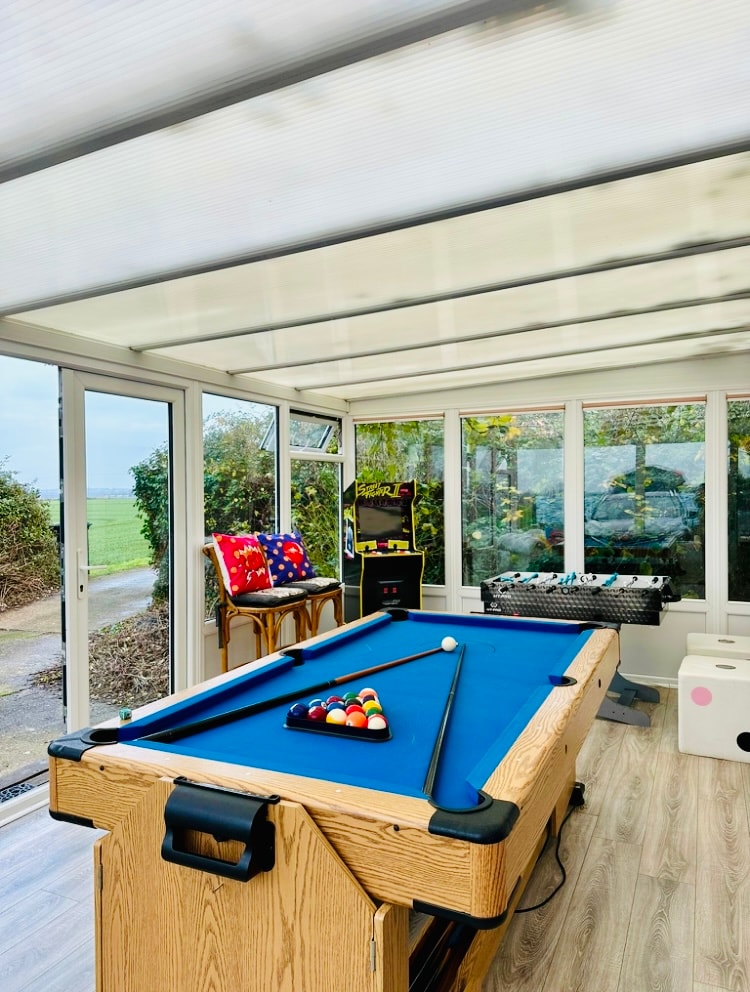 A bright games room features a wooden pool table with a blue felt surface and a triangle of colored balls positioned for play. Natural light floods in from multiple glass windows, illuminating arcade machines and additional game tables in the background.