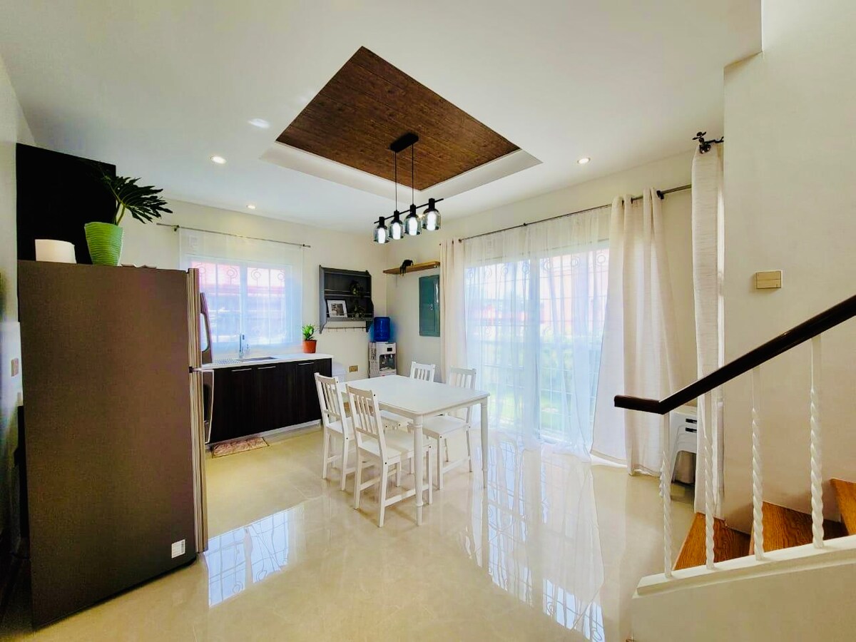 A modern dining area features a white table surrounded by four chairs, with soft natural light flowing through large windows. The adjoining kitchen includes dark cabinetry and counter space. A decorative plant is positioned on the counter, and wooden accents are visible above the dining area.