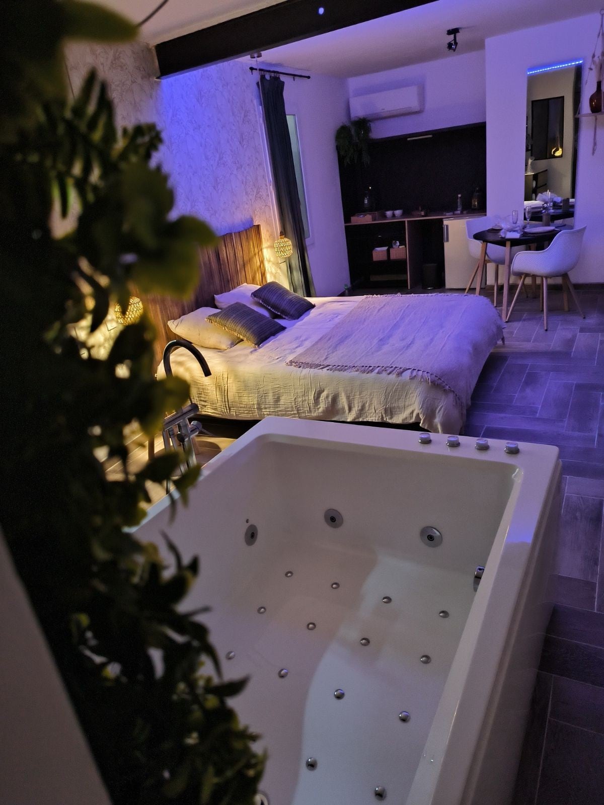 A modern suite features a spacious king-size bed with subtle bedding and decorative pillows. A balneo bathtub is positioned in the foreground, surrounded by soft lighting that highlights the room's relaxed ambiance. A small dining area with stylish chairs is visible in the background.
