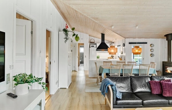 The open-plan living area features a spacious layout with light wooden beams above. A dining table is illuminated by two pendant lights, and a cozy seating area includes a dark sofa and a potted plant. The kitchen is equipped with modern appliances and a sleek design.