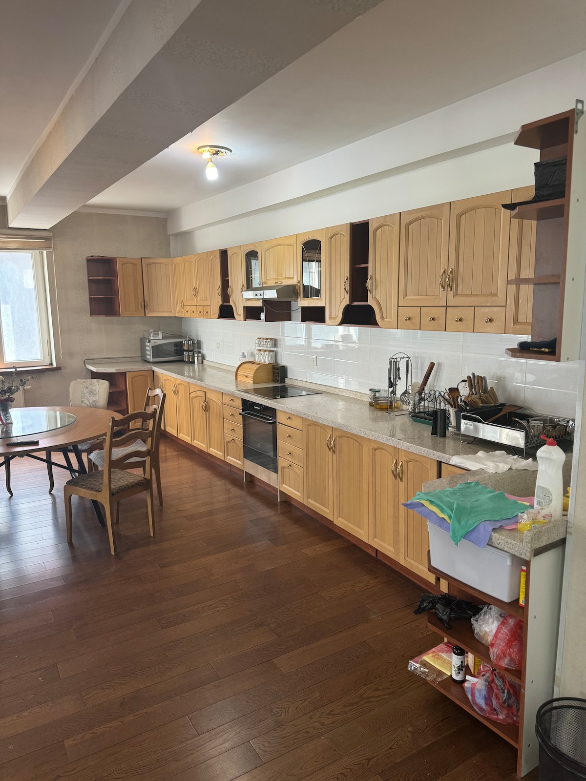 A spacious kitchen features wood cabinetry and a large countertop. Various kitchen appliances, including a microwave and oven, are visible. A dining table with four chairs is set for meals, and a window allows natural light to illuminate the space.