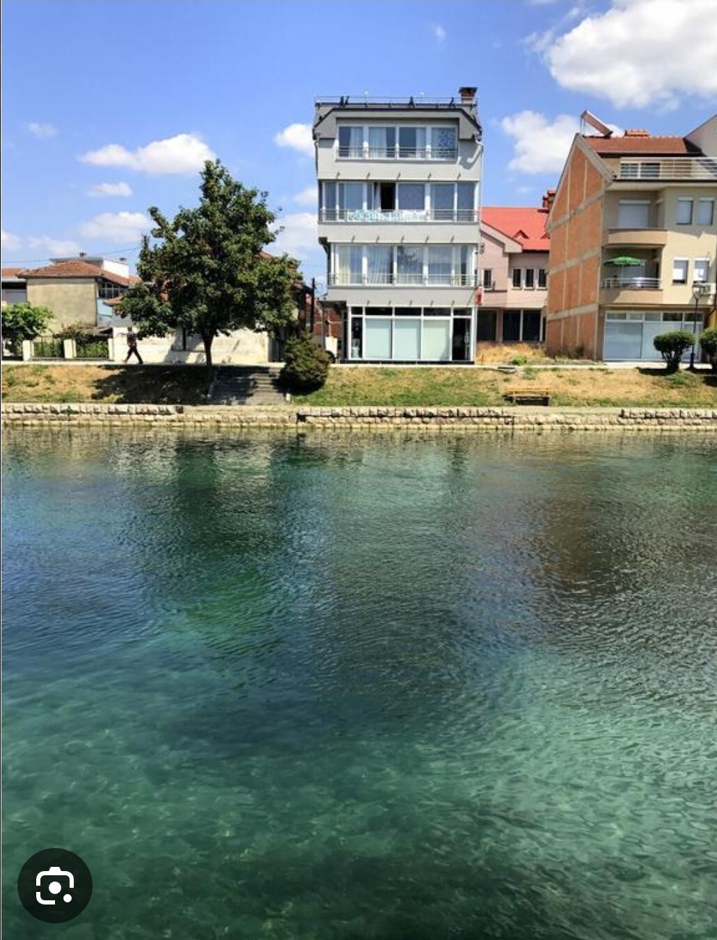A three-story building is situated along a clear body of water, reflecting the blue sky. The structure features large windows that provide views of the waterway. Nearby trees and a grassy area enhance the natural surroundings, contributing to a sense of tranquility.