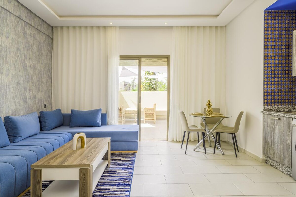 A well-lit living area features a spacious blue sectional sofa and a light wooden coffee table. A dining space with two chairs and a small table is positioned beside the large glass doors, which lead to an outdoor patio area. Soft curtains frame the windows.