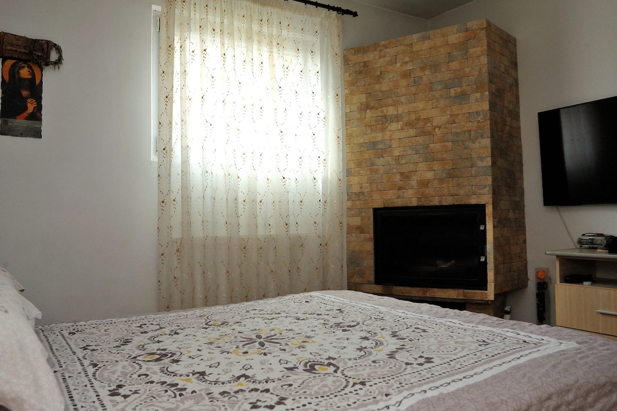 A spacious sleeping area is highlighted by a large bed covered with a decorative blanket. A textured stone feature wall complements the space, while a TV mounted on the wall provides entertainment options. Natural light filters through sheer curtains, adding brightness.