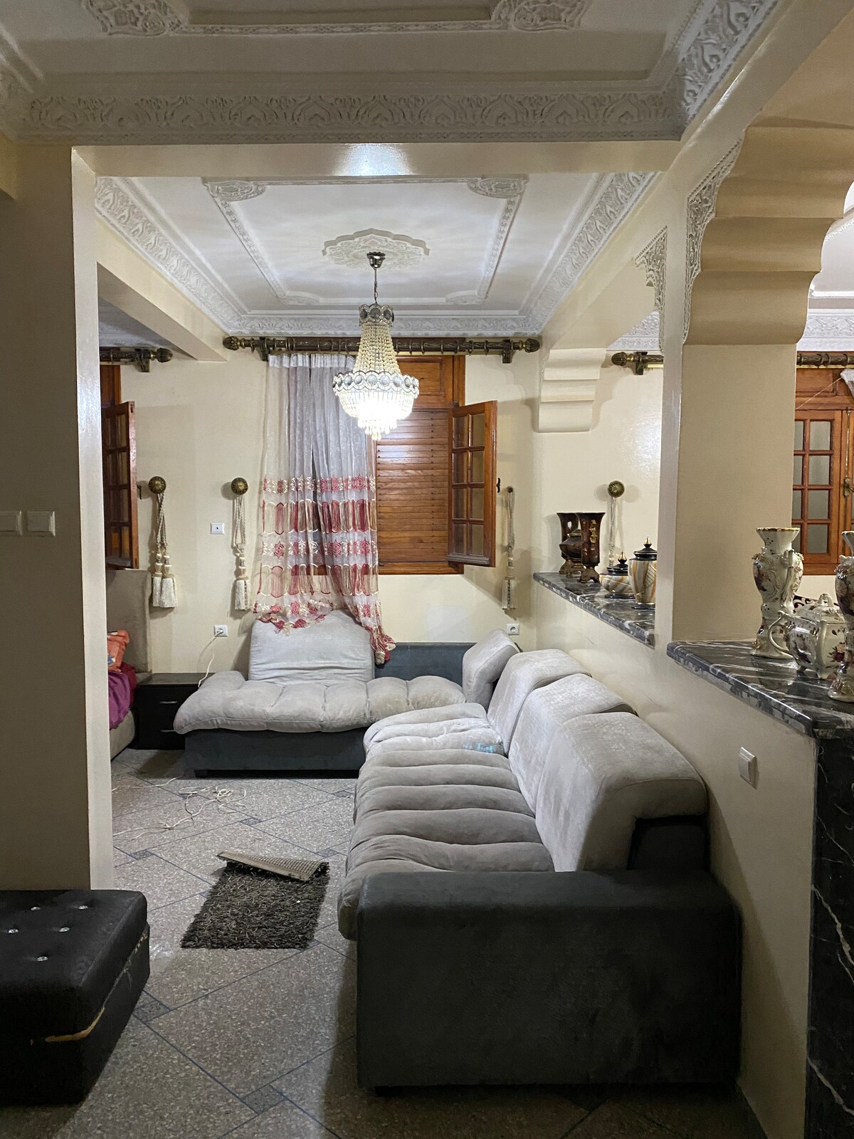 A spacious living area features a plush gray sectional sofa, complemented by a small black ottoman. Natural light enters through wooden windows adorned with fabric curtains. Decorative pieces are arranged on a contrasting marble surface, while intricate ceiling details enhance the ambiance.